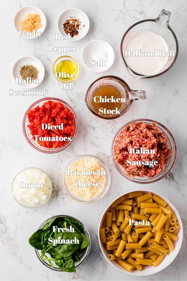 Ingredients for a pasta dish arranged on a marble countertop, including garlic, red pepper flakes, salt, Italian seasoning, olive oil, diced tomatoes, onion, fresh spinach, parmesan cheese, Italian sausage, pasta, chicken stock, and half-and-half.