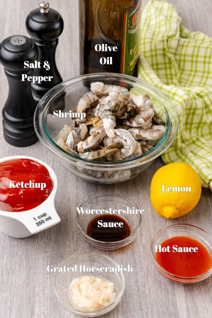 A kitchen counter displays ingredients for a shrimp cocktail: raw shrimp in a bowl, a bottle of olive oil, salt and pepper shakers, a lemon, grated horseradish, hot sauce, Worcestershire sauce, and a cup of ketchup. A green towel is in the background.