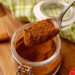 Spoon holding Apple Pie Spice Mix over glass jar with apples and cabbage in background on wooden surface