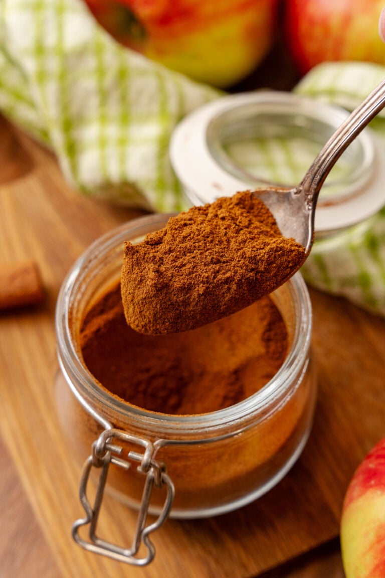Spoon holding Apple Pie Spice Mix over glass jar with apples and cabbage in background on wooden surface