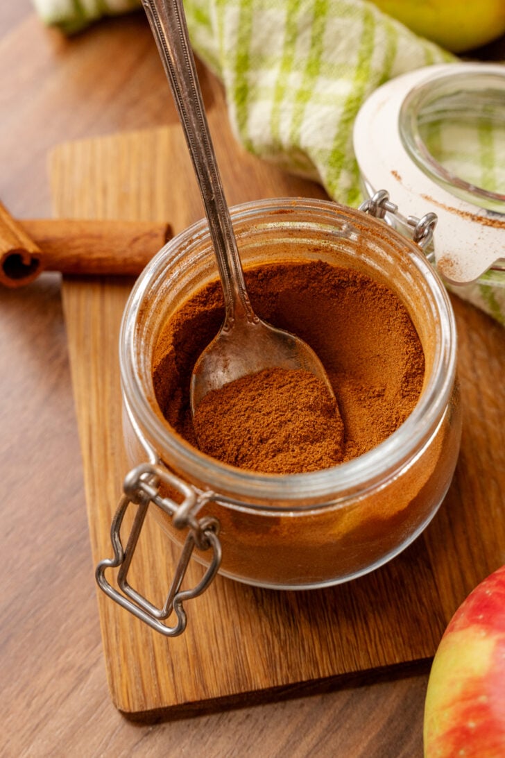 A metal spoon rests inside an open glass jar filled with ground cinnamon on a wooden cutting board. Nearby are a cinnamon stick, a green checkered cloth, and part of an apple.
