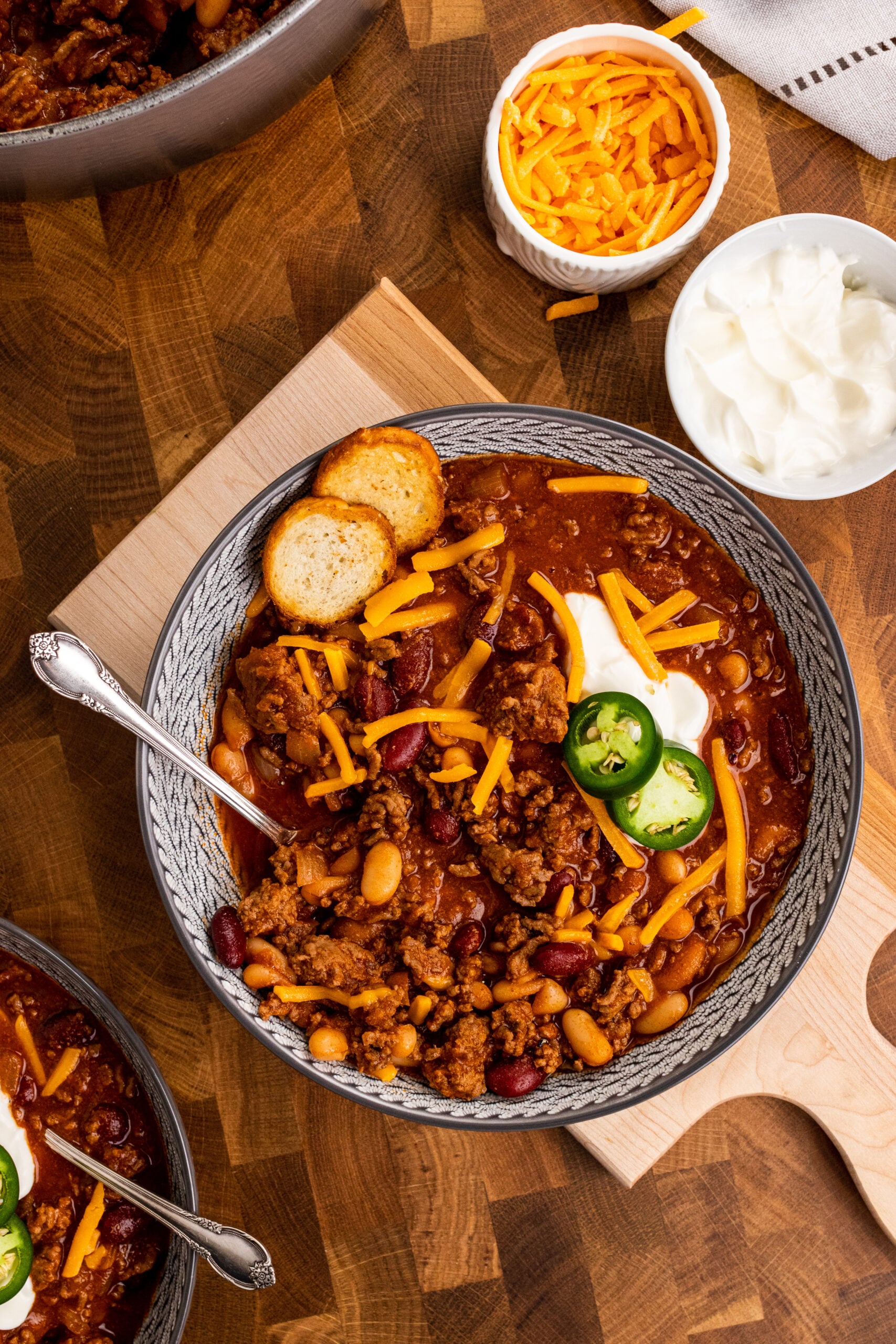 Homemade chili in gray bowl topped with shredded cheddar, sour cream, jalapeños, cornbread, and toppings for Quick Fall Pantry Meals for Bus