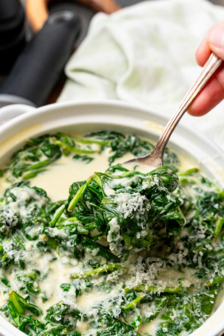 Creamed spinach topped with parmesan cheese in a white bowl served with a silver spoon.