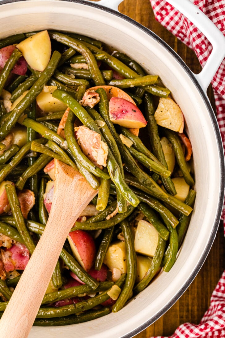 Southern green beans and potatoes with bacon cooked in a white dutch oven served with a wooden spoon.