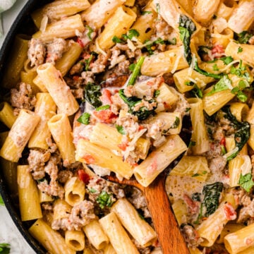 Italian sausage pasta with rigatoni, spinach, and diced tomatoes in a black skillet with wooden spoon