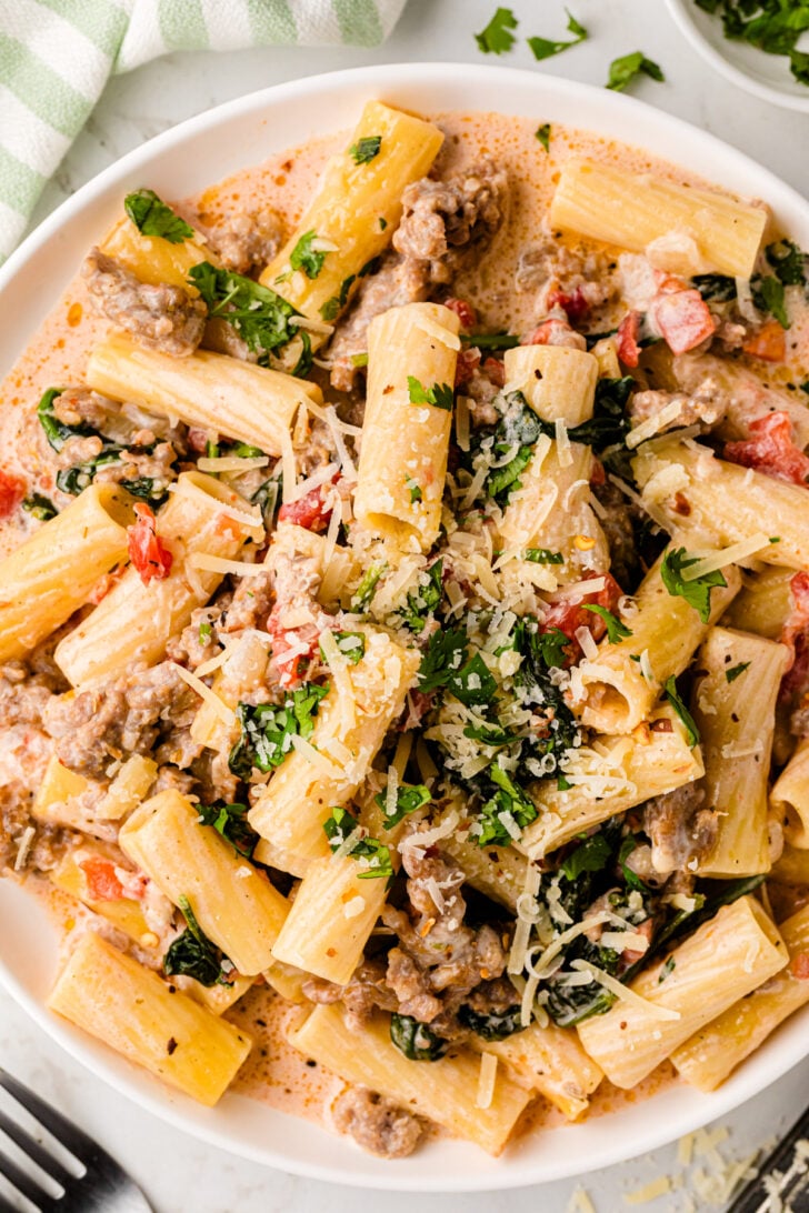A plate of creamy rigatoni pasta with ground meat, spinach, and diced tomatoes, garnished with grated cheese and fresh parsley. A striped napkin is partially visible in the corner.