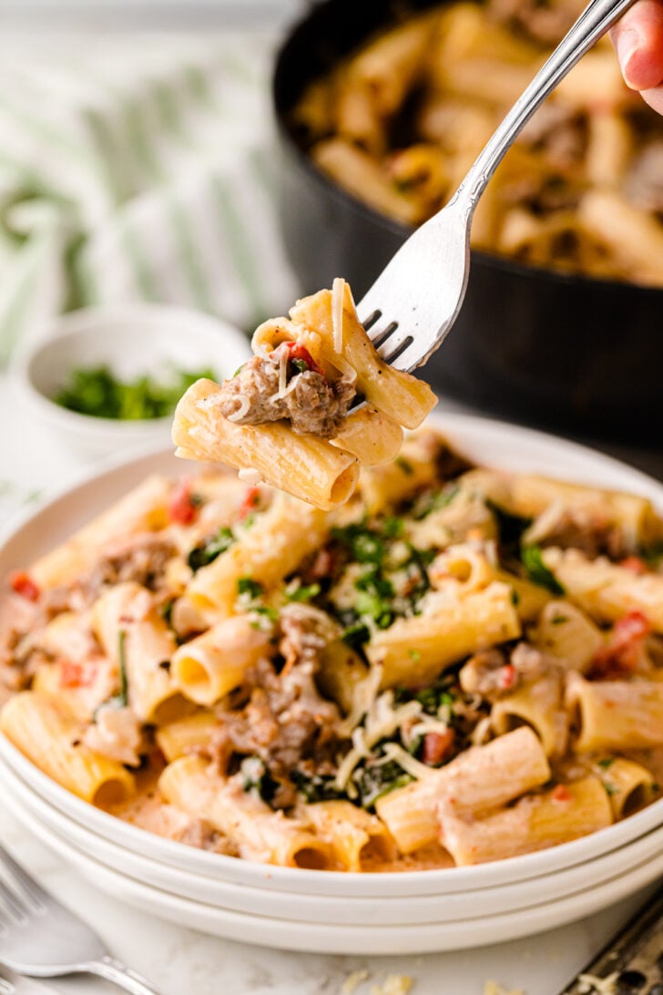 A fork lifting a bite of creamy pasta with sausage and herbs. The dish is served in a white bowl, with a black pot and a small dish of chopped herbs in the background. The pasta is topped with grated cheese and garnished with greens.