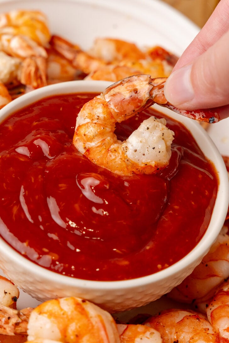 A hand dipping a cooked shrimp into a bowl of red cocktail sauce, surrounded by more shrimp on a white plate.