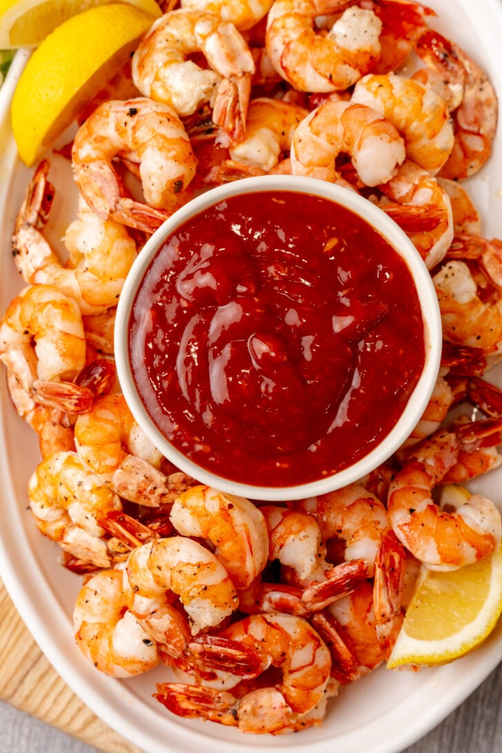 A plate of cooked shrimp arranged around a bowl of red cocktail sauce. Lemon wedges are placed on the side of the plate for garnish.