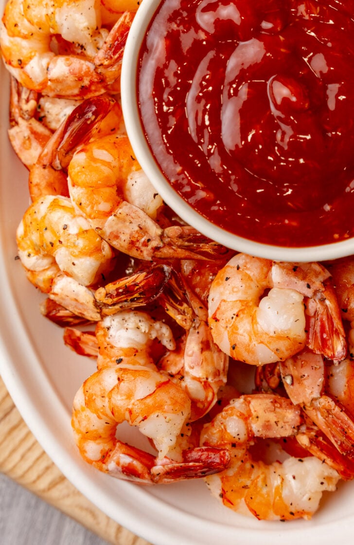 A plate of seasoned, cooked shrimp surrounds a bowl of red cocktail sauce. The shrimp are arranged in a circular pattern, displaying a mix of orange and white colors. The cocktail sauce appears thick and glossy.