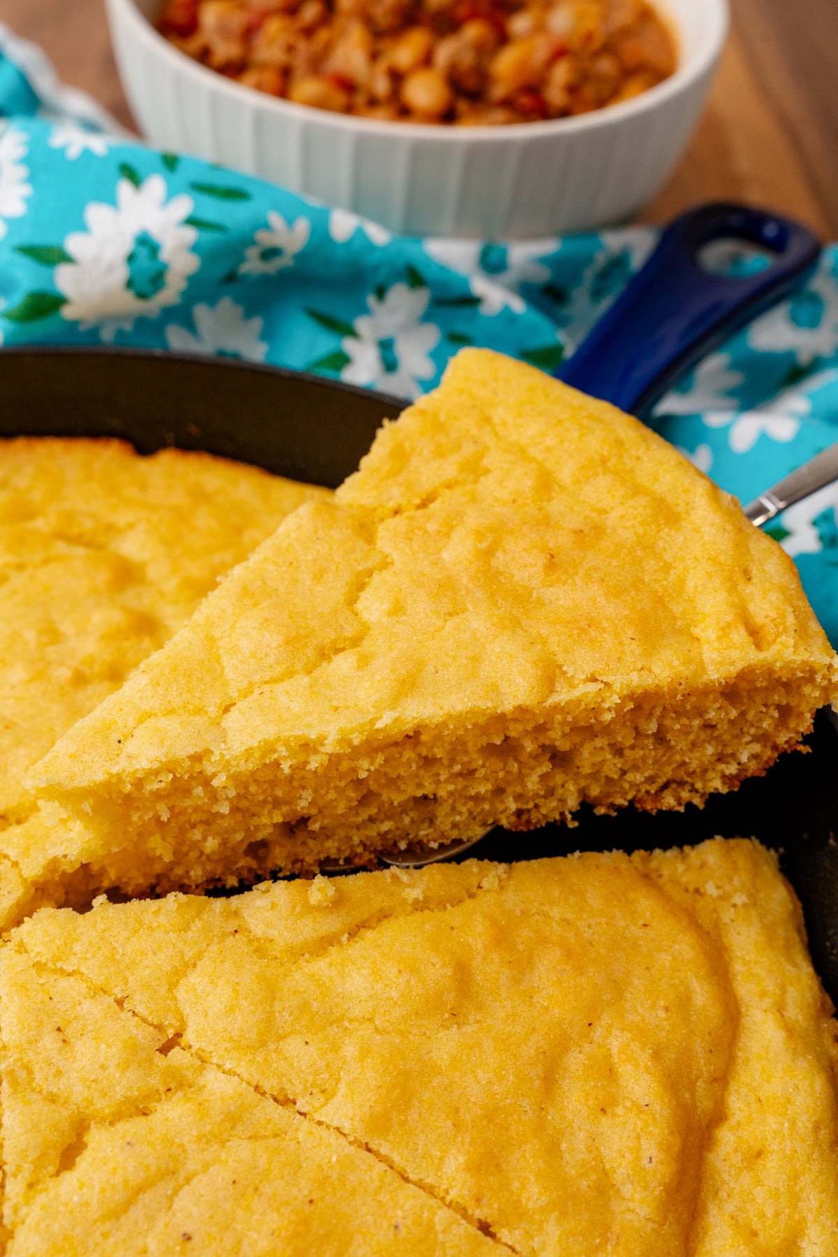 Slice of golden southern cornbread 6 on a knife with turquoise floral cloth and white bowl in background