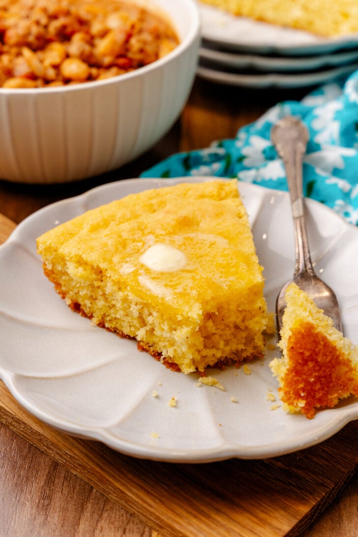 Slice of old-fashioned Southern cornbread with butter on a white plate served with a silver spoon.