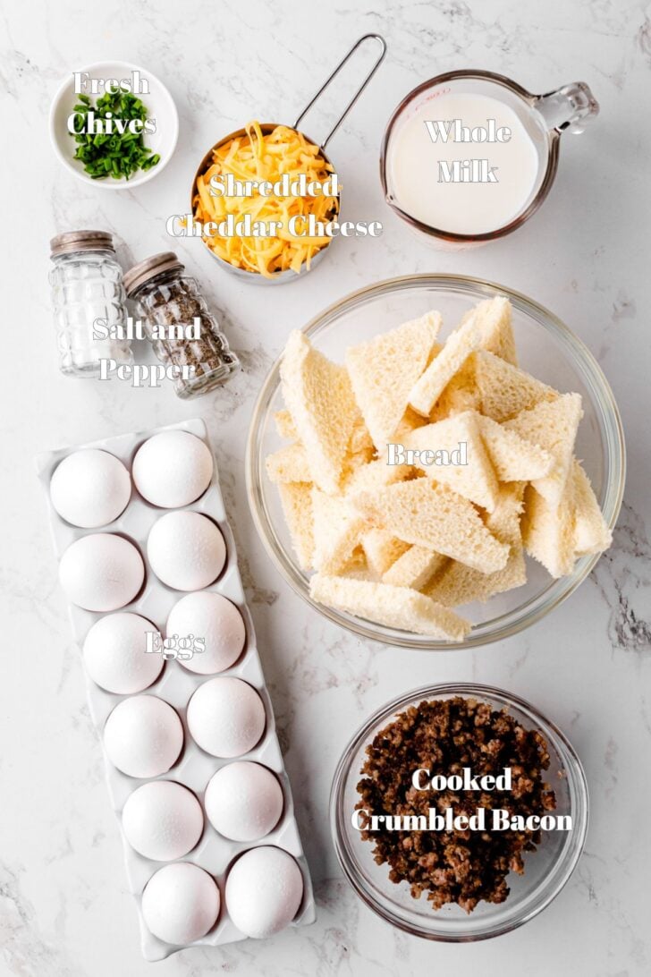 Ingredients on a marble surface: fresh chives, shredded cheddar cheese, whole milk, sliced bread, salt and pepper shakers, a dozen eggs, and a bowl of cooked crumbled bacon.