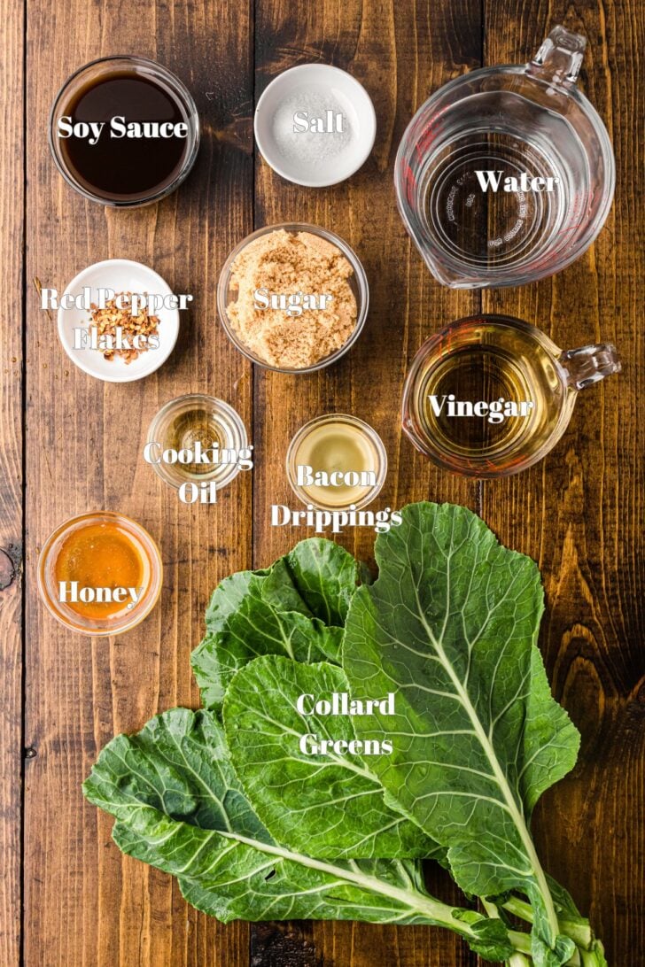 Ingredients for a recipe are laid out on a wooden surface. Items include soy sauce, salt, red pepper flakes, sugar, water, vinegar, cooking oil, bacon drippings, honey, and fresh collard greens.
