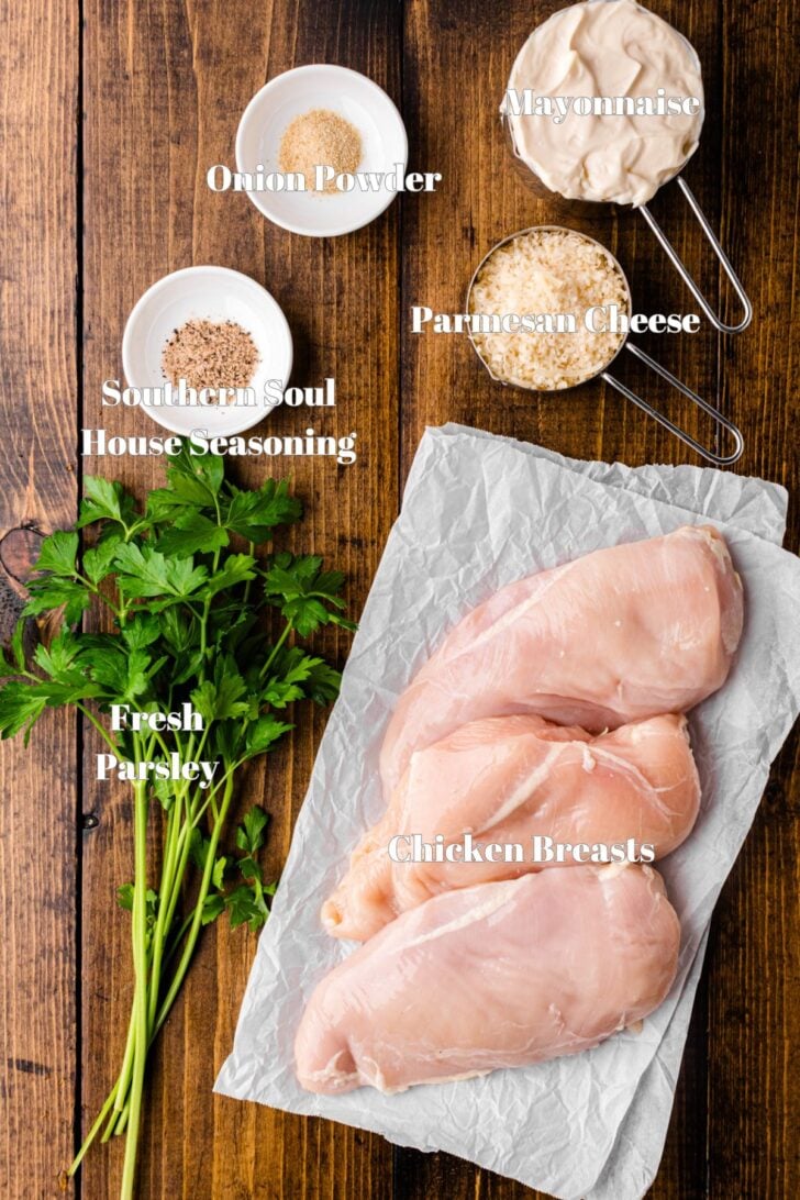 Top-down view of raw chicken breasts on parchment paper, surrounded by ingredients: fresh parsley, Southern Soul House Seasoning, onion powder, parmesan cheese, and mayonnaise. All items are labeled on a wooden surface.