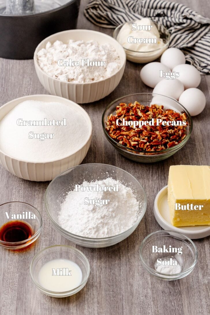 A wooden table displays various baking ingredients: bowls of cake flour, granulated sugar, chopped pecans, powdered sugar, and baking soda. Nearby are eggs, butter, sour cream, vanilla extract, and milk, with a striped cloth in the background.