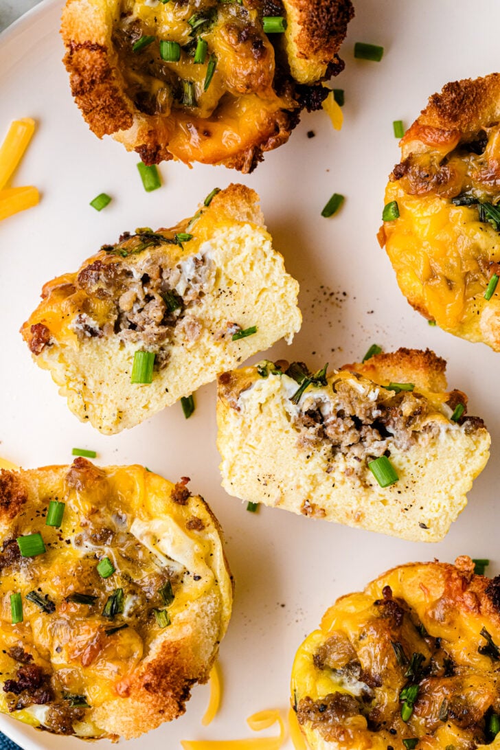 A plate of savory breakfast muffins topped with melted cheese and green herbs. The muffins have a golden-brown crust. In the background, there are eggs, spices, and chopped herbs on a table.