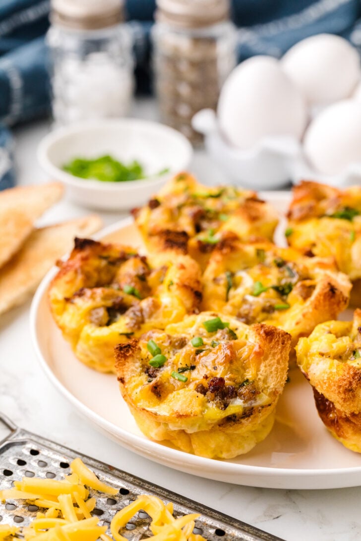 A plate of savory breakfast muffins topped with melted cheese and green herbs. The muffins have a golden-brown crust. In the background, there are eggs, spices, and chopped herbs on a table. A cheese grater with shredded cheese is in the foreground.