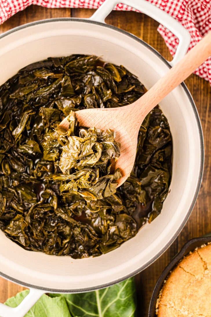 A pot of cooked collard greens with a wooden spoon lifting some out. A red checkered cloth and a piece of cornbread are in the background.