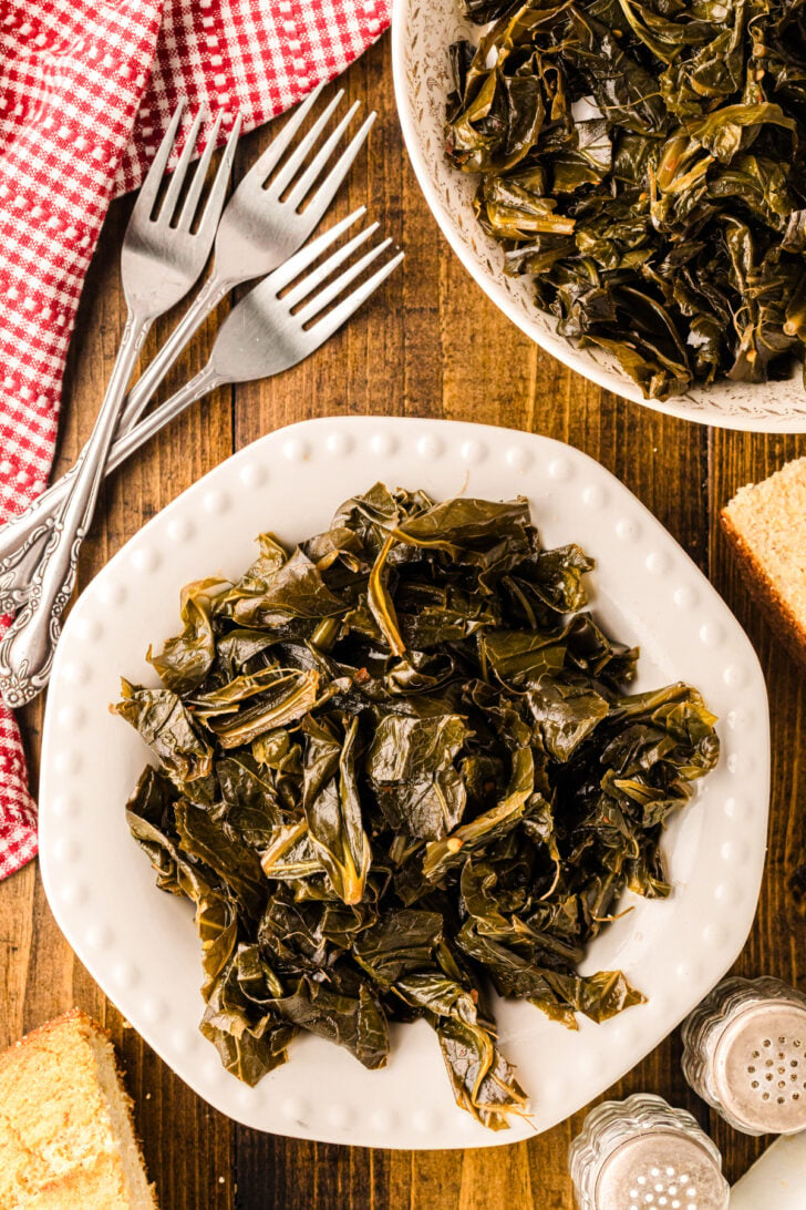A bowl filled with cooked collard greens, with a spoon resting inside. The dish is set on a wooden table next to a red and white checkered cloth.
