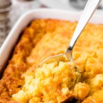 A spoon lifts a creamy serving of Classic Cornbread Pudding With Cheese from a white baking dish, showing the golden-brown top and tender in
