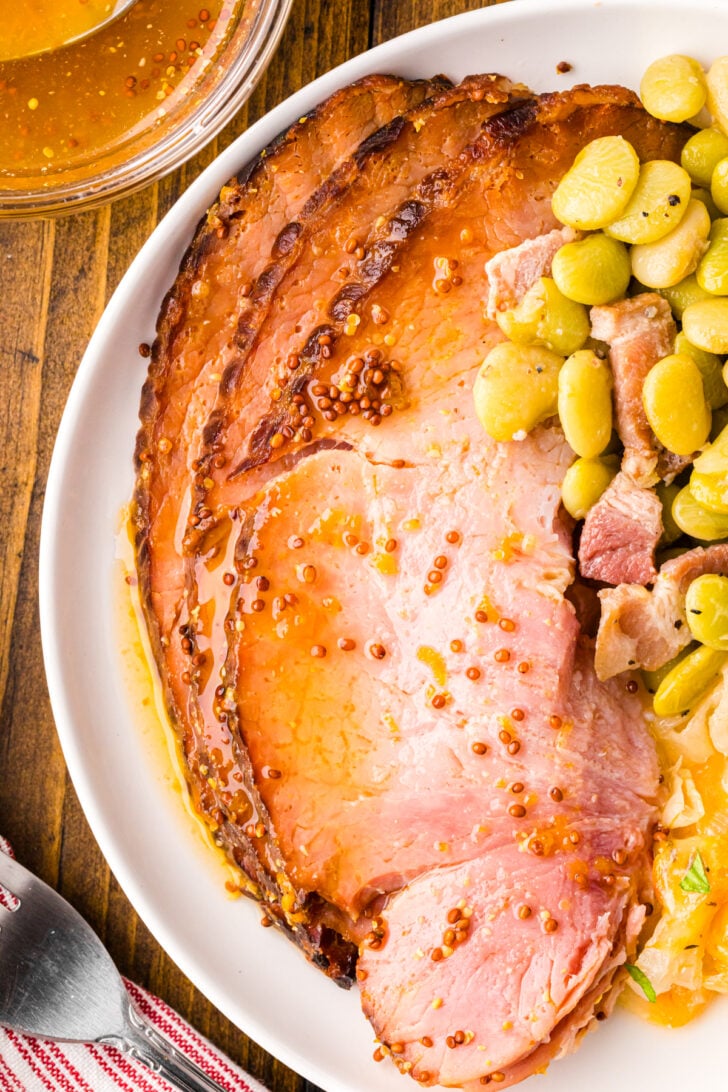 A plate featuring glazed ham slices is garnished with mustard seeds, accompanied by cooked lima beans and chunks of bacon. The dish rests on a wooden table, next to a red-striped napkin and a small bowl of sauce.