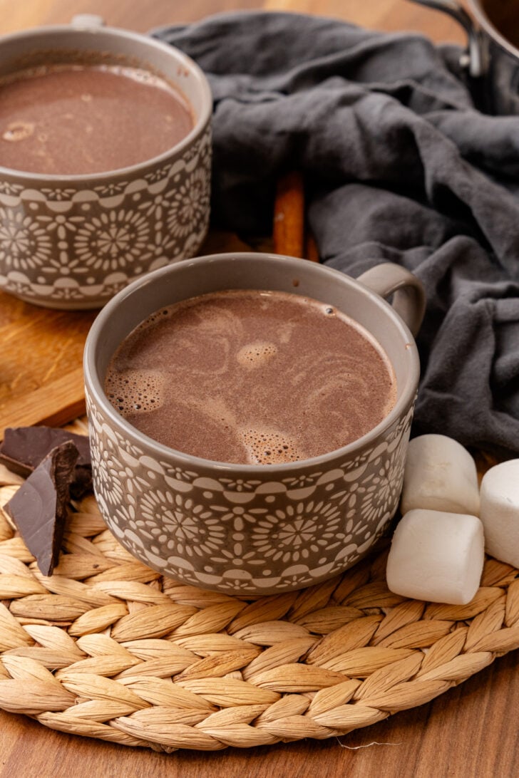 Two gray mugs filled with hot chocolate sit on a woven mat. The mugs have a floral pattern. Nearby, there are marshmallows, dark chocolate pieces, and a dark cloth. The scene suggests a cozy and warm atmosphere.