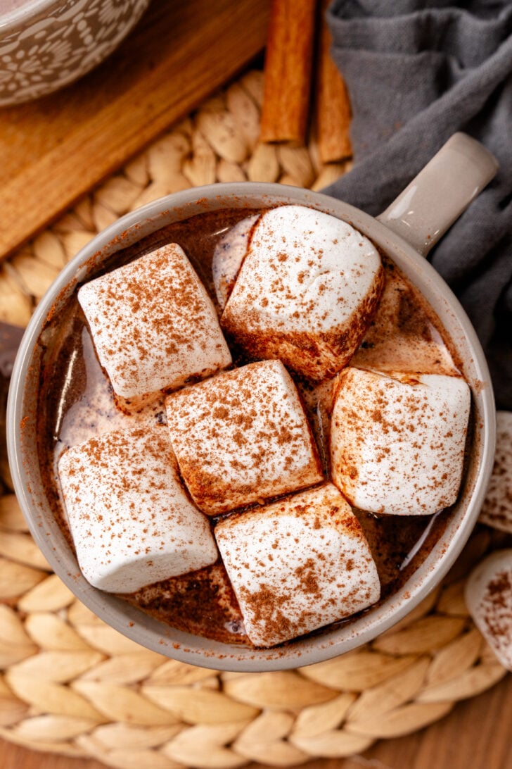 A mug of hot chocolate topped with large marshmallows, sprinkled with cocoa powder. It's placed on a woven placemat, with cinnamon sticks nearby. A gray cloth is partially visible to the side.