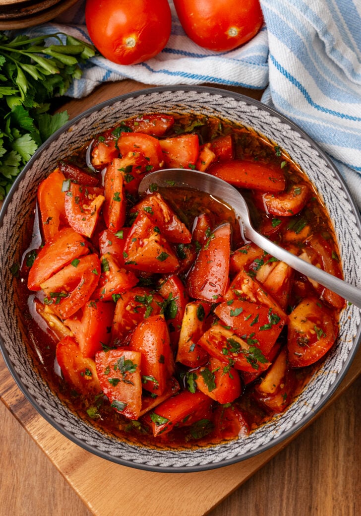Marinated tomatoes 4 served in decorative bowl with fresh parsley garnish, spoon, and whole tomatoes visible in background on wooden surface