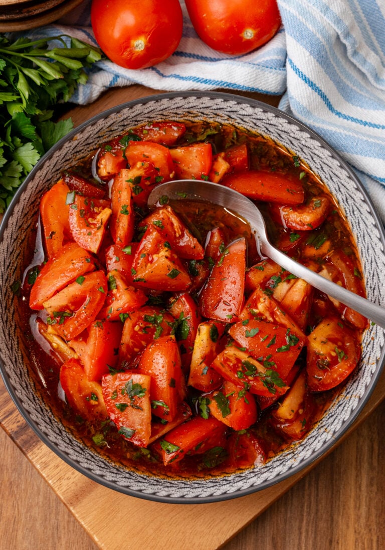 Marinated tomatoes 4 served in decorative bowl with fresh parsley garnish, spoon, and whole tomatoes visible in background on wooden surface