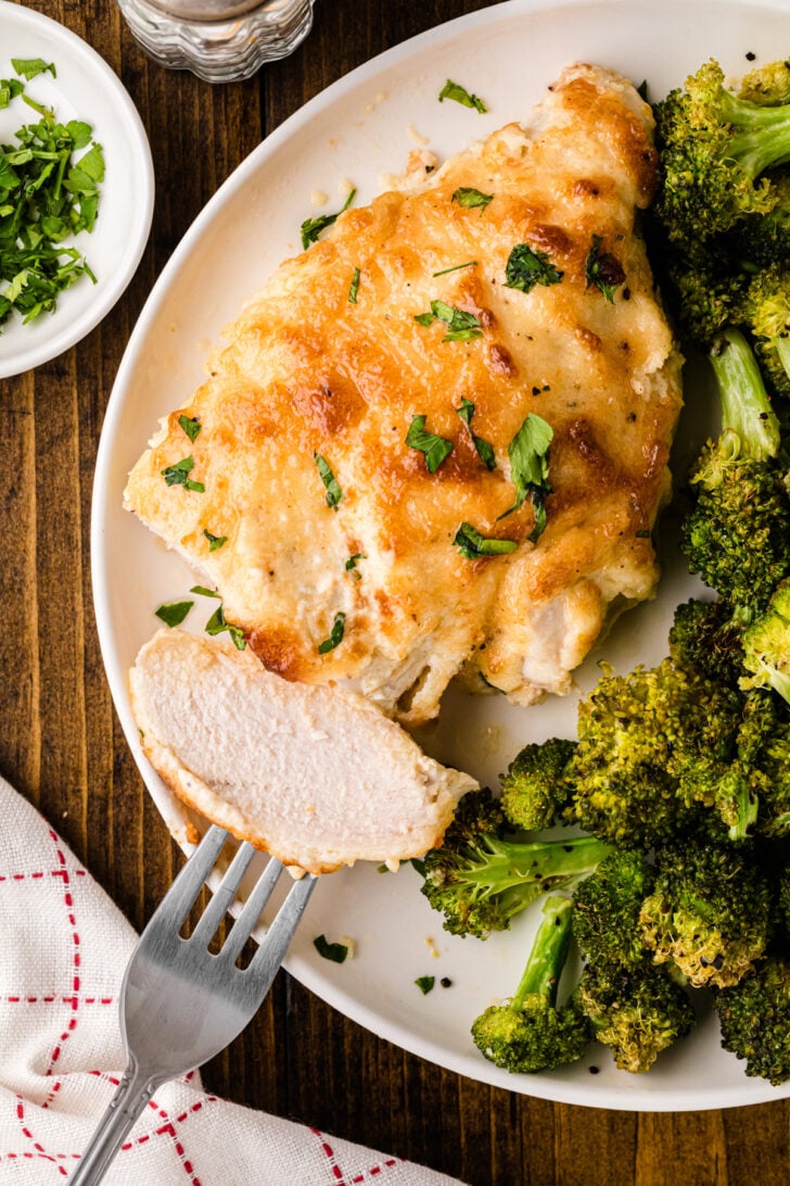 Melt In Your Mouth Chicken breast on white plate with roasted broccoli, garnished with fresh parsley, fork beside, overhead view