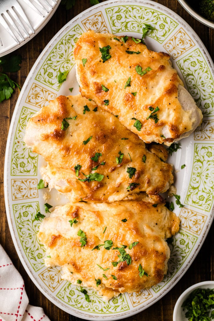 Three golden-brown baked chicken breasts garnished with chopped parsley, served on an ornate oval platter. The dish is set on a wooden table, with a striped cloth and small bowl of herbs nearby.