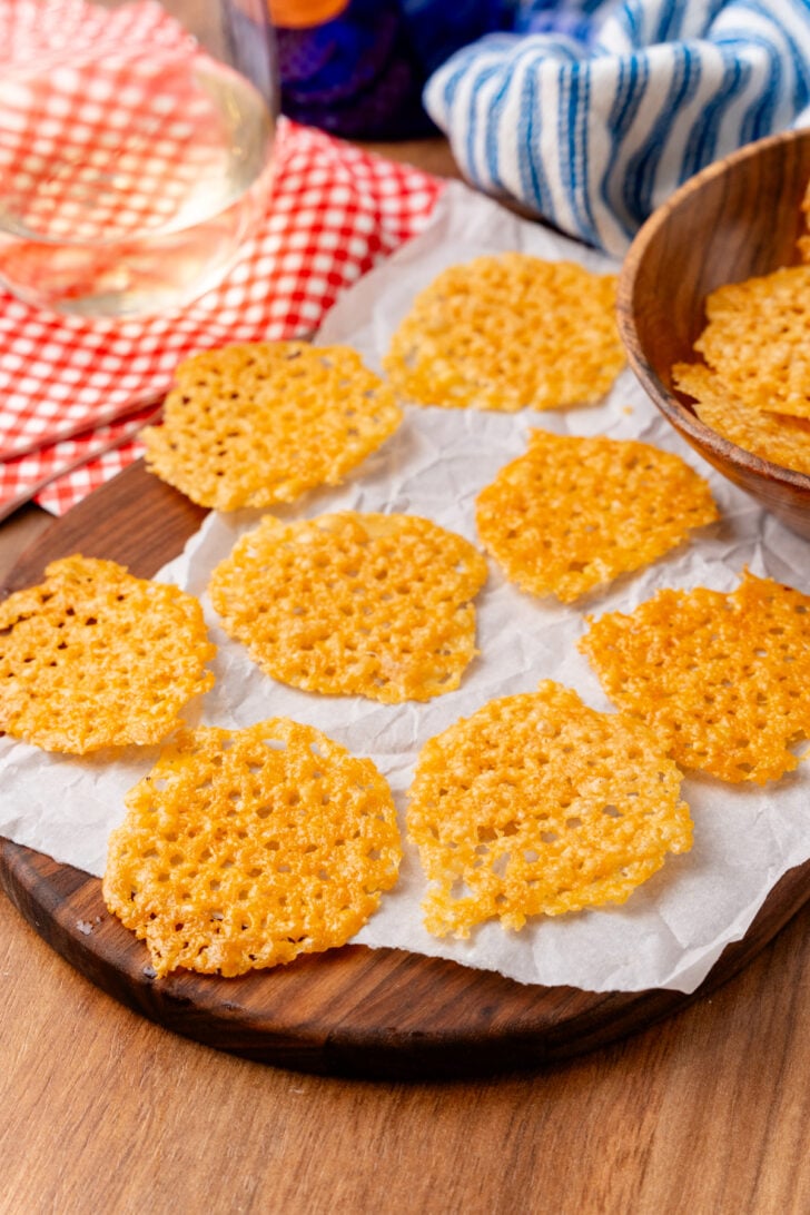 Baked parmesan cheese crisps on parchment paper and a serving board served with a glass of wine.