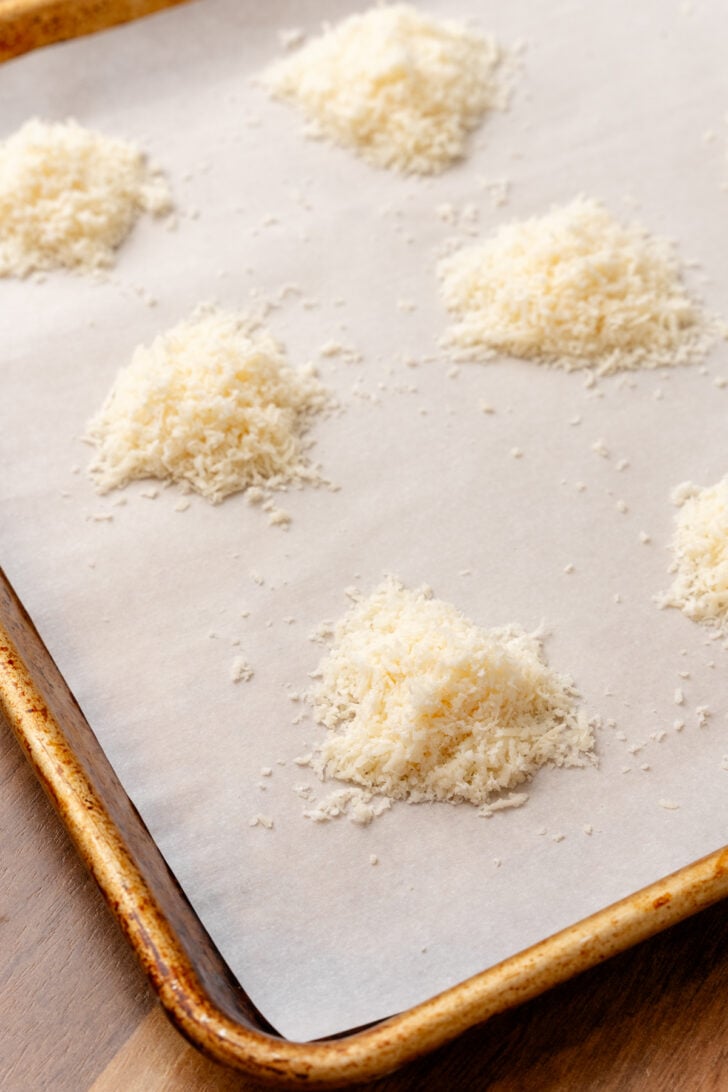Parchment lined baking sheet with mounds of grated parmesan cheese for baking.
