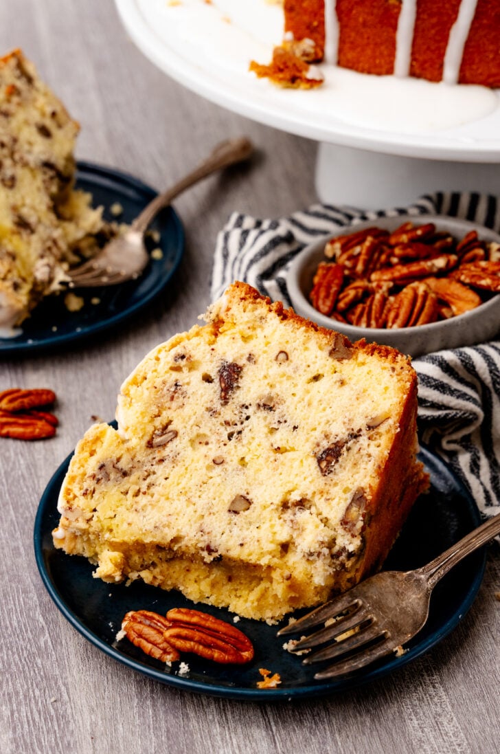 A slice of pecan cake with visible nuts on a blue plate, accompanied by a fork and whole pecans. The background shows a striped cloth and another plate with a cake slice. A partially visible whole cake is on a stand in the background.