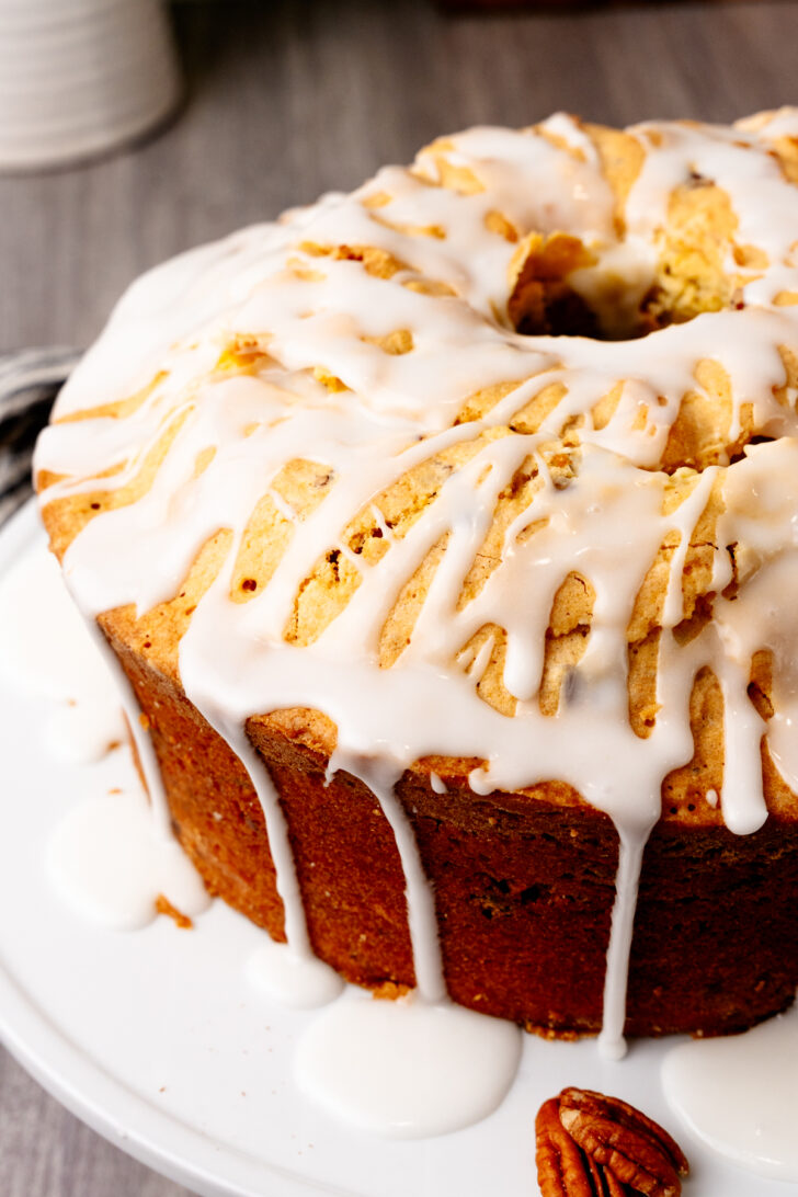A bundt cake topped with glossy white icing, displayed on a white plate. The cake has a light brown color with pieces of pecans scattered around it, resting on a wooden surface.