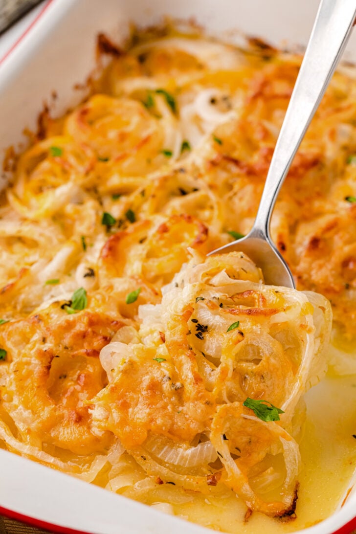 A close-up of a baked dish featuring golden-brown, cheese-covered onions. The onions are thinly sliced and garnished with herbs. A spoon is lifting a portion, revealing a creamy sauce underneath in a white and red baking dish.