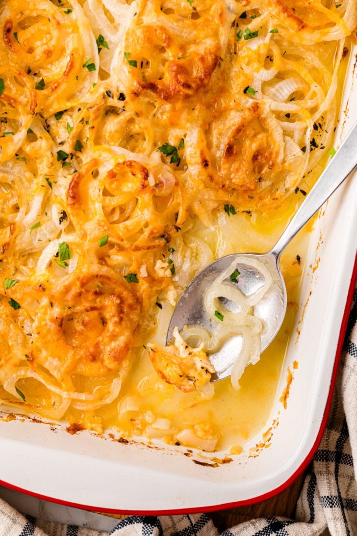 A baking dish filled with creamy, cheesy onions au gratin, topped with fresh herbs. A spoon rests on the right, serving a portion. The dish is placed on a checkered cloth.