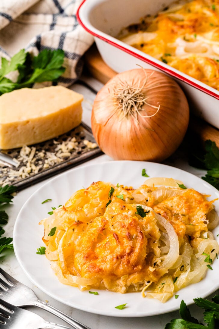 A plate of cheesy baked potatoes topped with onions and garnished with parsley. In the background, there's a block of cheese, a whole onion, and a baking dish with more of the potato dish. Silverware and a checkered cloth are nearby.