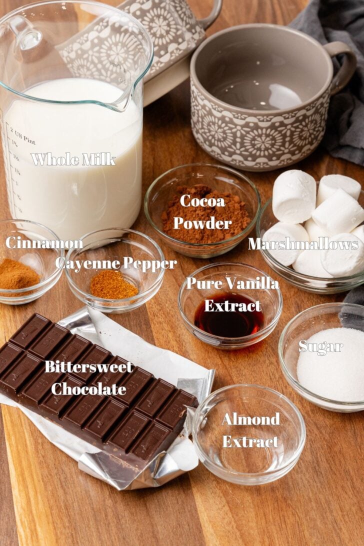 A wooden table displays ingredients for hot chocolate: a measuring cup of whole milk, a patterned mug, bowls of cocoa powder, cinnamon, cayenne pepper, pure vanilla extract, marshmallows, sugar, a bar of bittersweet chocolate, and almond extract.