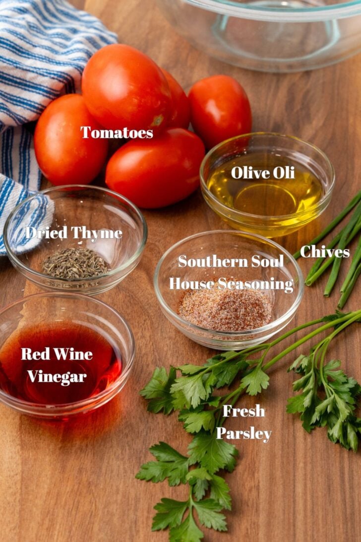 A wooden table with fresh tomatoes, olive oil, dried thyme, red wine vinegar, Southern Soul house seasoning, chives, and fresh parsley arranged alongside a striped cloth napkin and an empty glass bowl.