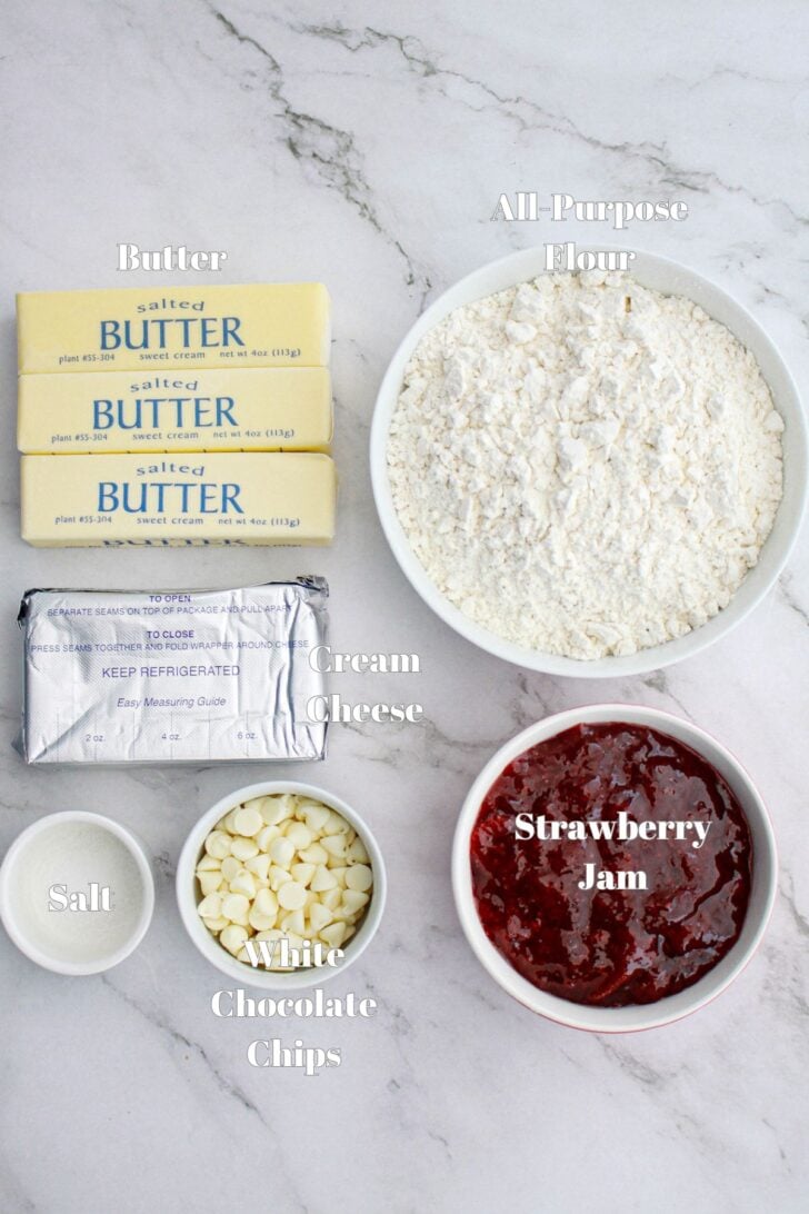 Ingredients on a marble surface include: three sticks of butter, a block of cream cheese, a bowl of all-purpose flour, a bowl of strawberry jam, a bowl of white chocolate chips, and a small bowl of salt.