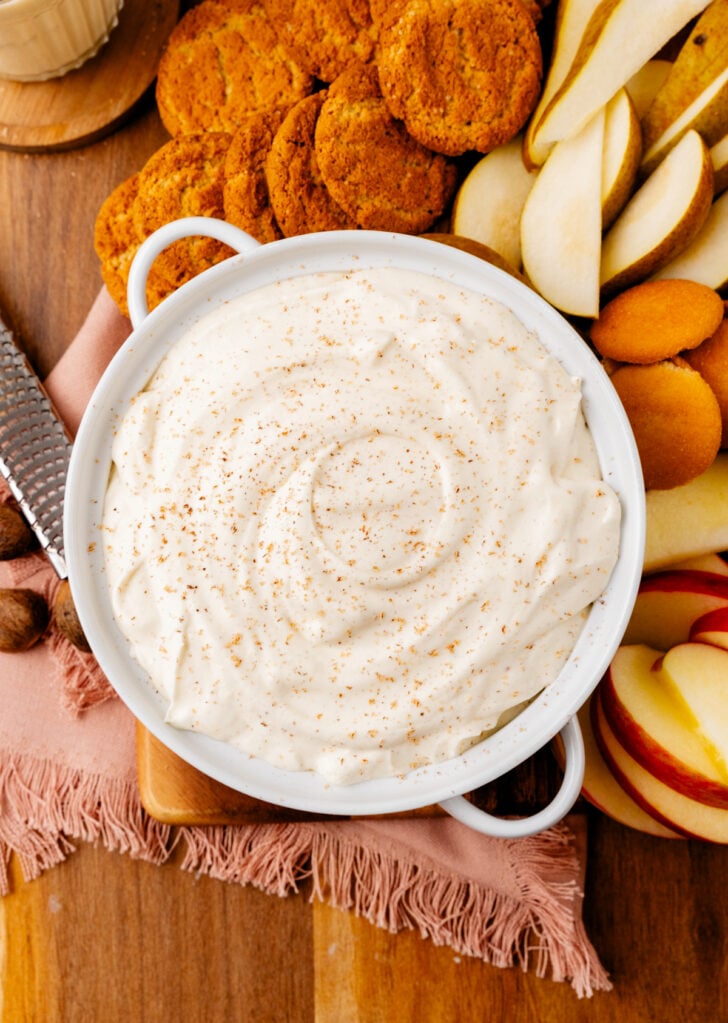 White bowl of Eggnog Cheesecake Dip served with apple slices, cookes and gingersnaps.