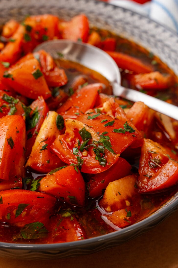 A bowl of fresh tomato salad with herbs and a tangy dressing. The salad features vibrant red tomato wedges, finely chopped herbs, and is glistening with the dressing. A spoon is placed in the bowl, ready for serving.