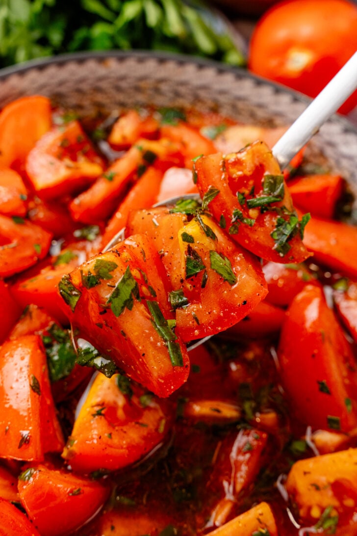 Close-up of a bowl of vibrant, juicy tomato salad with chopped parsley and a vinaigrette dressing. A spoon is scooping up a portion, highlighting the fresh ingredients and herbs.