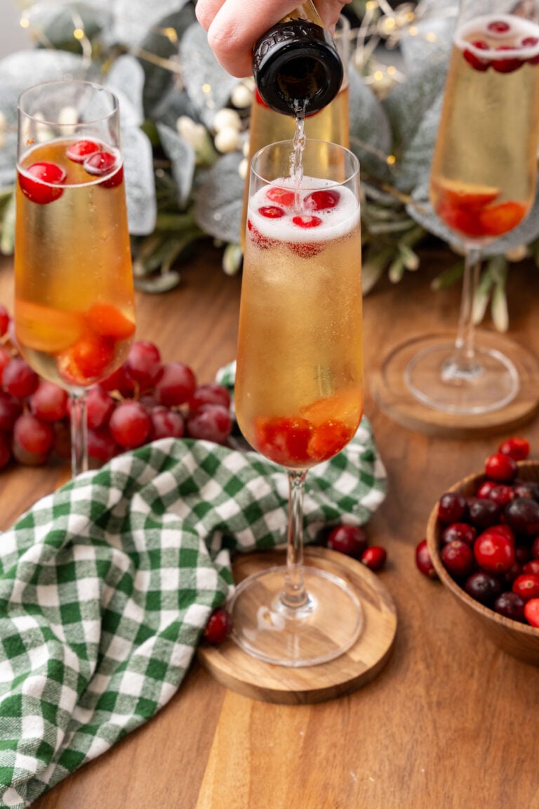 Pouring champagne into a mistletoe mimosa 11 cocktail garnished with red berries in a flute glass on a wooden table