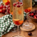 Mistletoe mimosa 4 in champagne flute with red berries and ice, wooden coaster, bottle and additional glasses on wooden table