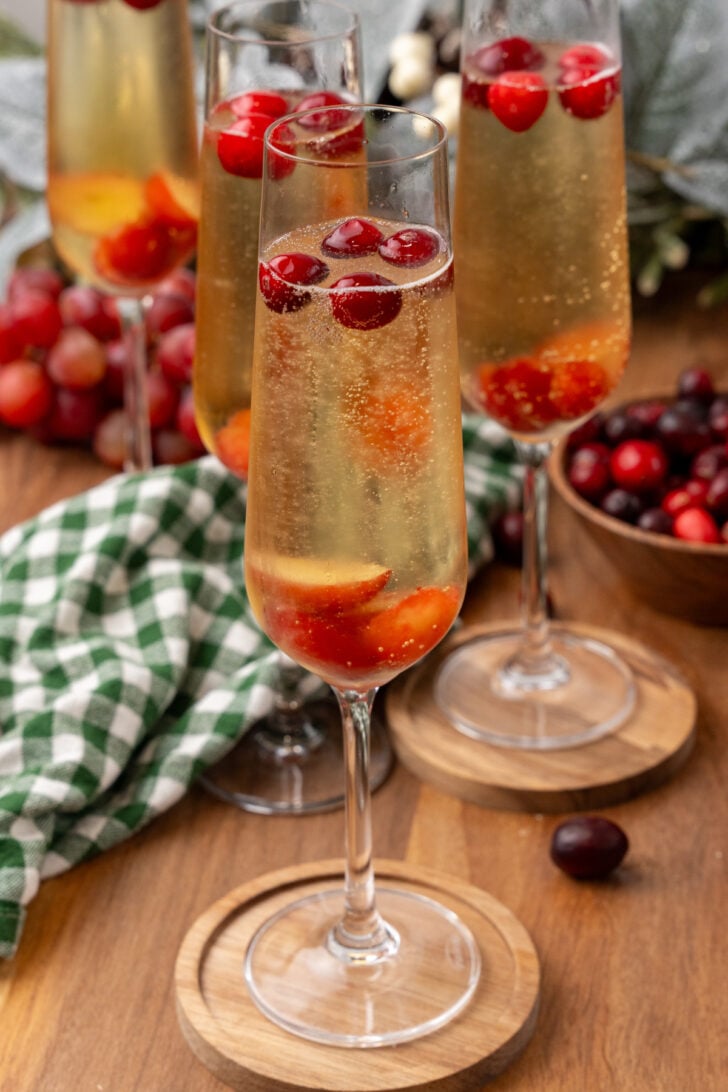 Mistletoe mimosa 4 in champagne flute with red berries and ice, wooden coaster, bottle and additional glasses on wooden table