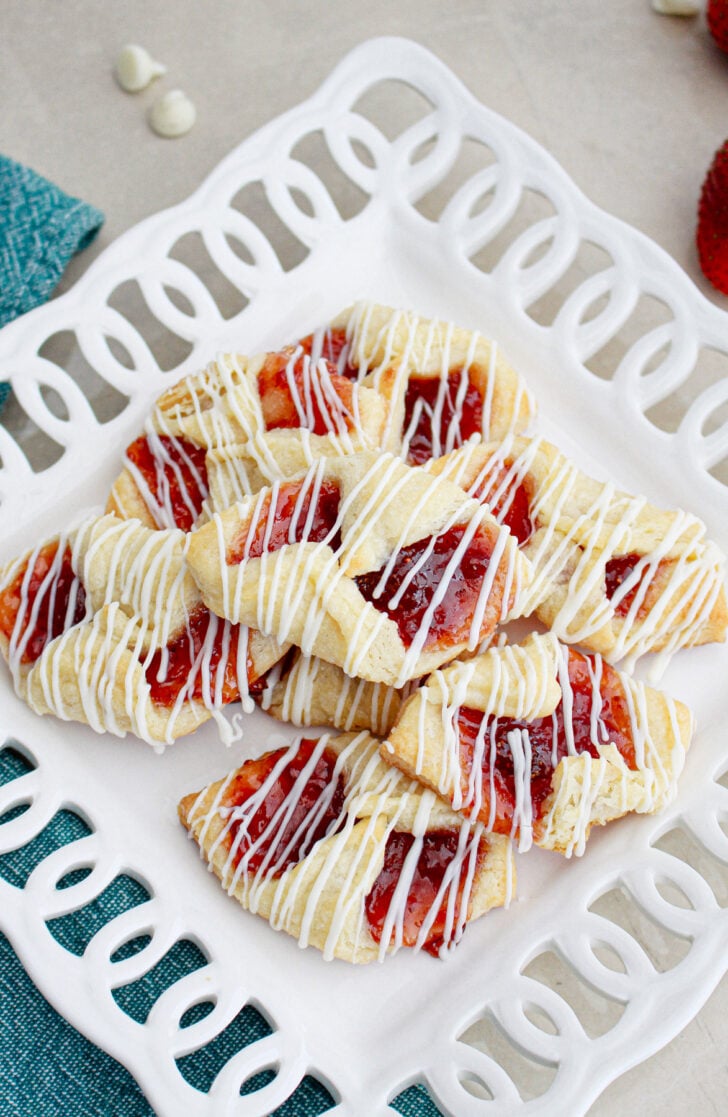 A white decorative plate holds several pastries filled with red fruit jam, drizzled with white icing. The plate is set on a turquoise cloth, and a few small white chocolate chips are scattered nearby.