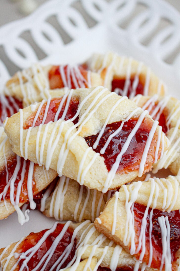 A plate of pastries with a golden-brown crust, filled with bright red jam and drizzled with white icing. They are arranged in a visually appealing stack on a patterned white plate.